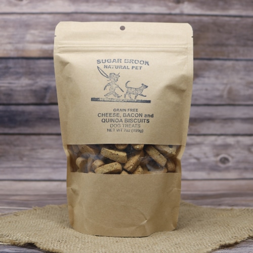Sugar Brook grain-free Cheese, Bacon, and Quinoa Biscuits Dog Treats in a kraft stand-up pouch, displayed on a burlap mat against a wooden backdrop.