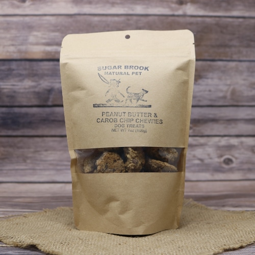 Sugar Brook Natural Pet Peanut Butter & Carob Chip Chewies Dog Treats in a kraft paper pouch with a see-through window, on a burlap mat with wooden background.