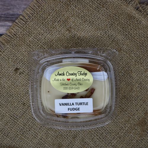 Clear plastic Ashery bowl with yellow sticker label on the plastic lid, filled with vanilla turtle fudge, sitting on a burlap material with wood background