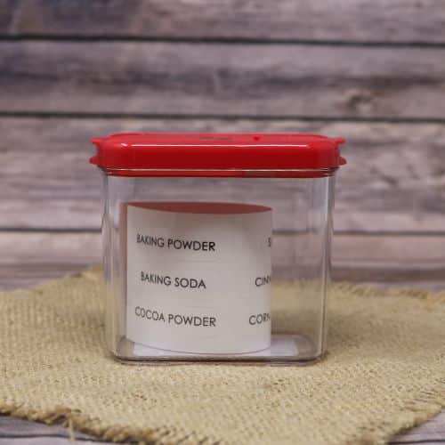Clear plastic container with red lid that has a piece of paper with a list of ingredients, sitting on a burlap material with a wood background