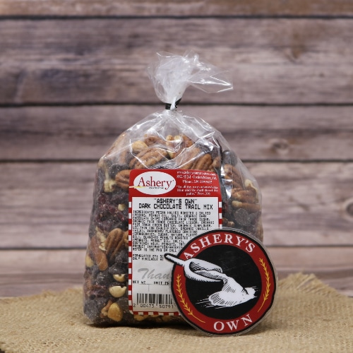 A bag of Ashery's Own Dark Chocolate Trail Mix, filled with nuts and chocolate pieces, displayed on a woven straw mat with a wood grain background.
