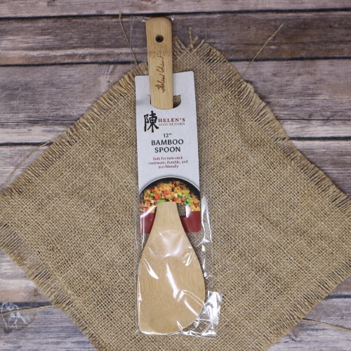 A Helen's Asian Kitchen bamboo spoon, wrapped in plastic packaging, lies on a rustic straw mat with a wooden backdrop.