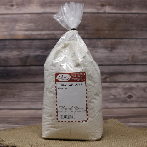 A bag of Ashery organic barley flour tied at the top, standing on a woven straw mat against a wooden plank background.