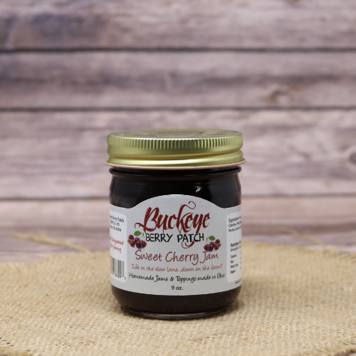 A jar of Buckeye Berry Patch Sweet Cherry Jam with a golden lid, centered on a straw mat with a wooden plank background."