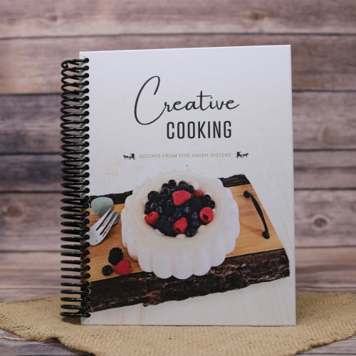 Creative Cooking cookbook with a dessert on the cover, placed on a burlap mat with a rustic wooden background.