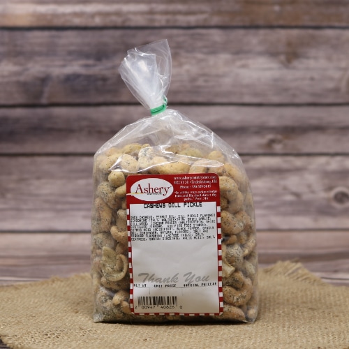 A sealed bag of Ashery dill pickle-flavored cashews on a textured straw mat, with a softly focused wooden background.