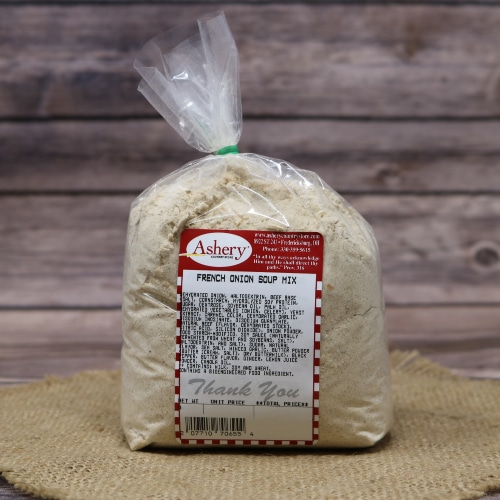 A sealed bag of Ashery brand French Onion Soup Mix on a circular straw mat, with a rustic wooden background. The label on the bag includes product and nutritional information