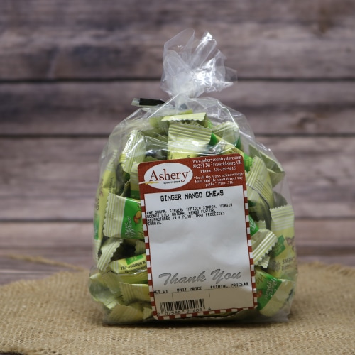 A bag of Ashery Ginger Mango Chews candy, with a mix of green and white wrappers, tied off with a twist and placed on a straw mat with a wooden backdrop.