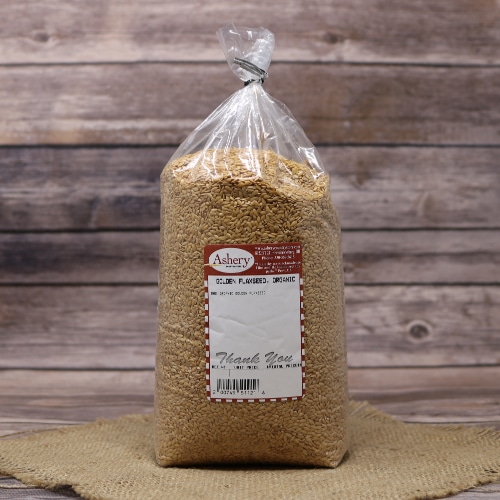 A tall, transparent bag of Ashery golden flaxseed placed on a circular straw mat with a soft-focus wooden background.