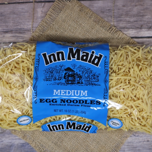 A package of Inn Maid Medium Egg Noodles against a backdrop of loose noodles on a straw mat, with a rustic wood background.