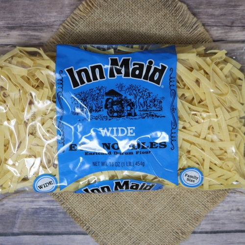 A bag of Inn-Maid Wide Noodles placed atop a straw mat, with loose noodles scattered around, against a wood-patterned backdrop