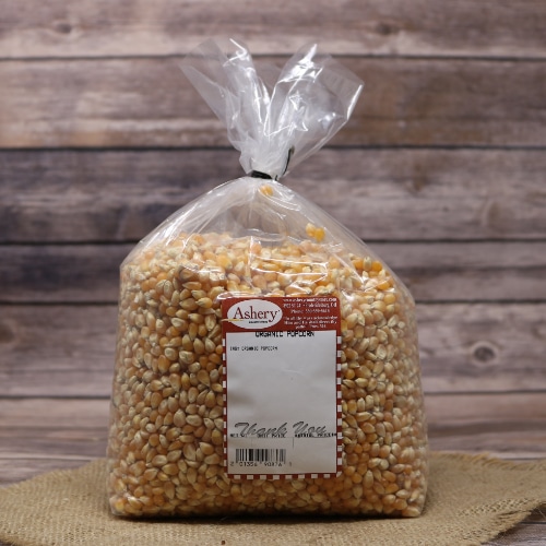 Large clear bag of Asher's Organic Ladyfinger Popcorn kernels with a white and red label, on a burlap surface with a wooden background.