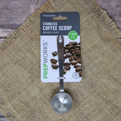 A Progressive Prepworks stainless steel coffee scoop with a 1-tablespoon measure, attached to a card with coffee beans graphic, on a burlap surface.
