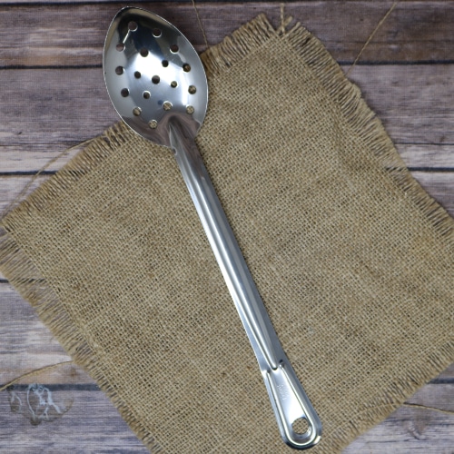 A stainless steel perforated spoon laid out on a hessian burlap mat, with a wooden plank background providing a natural contrast.