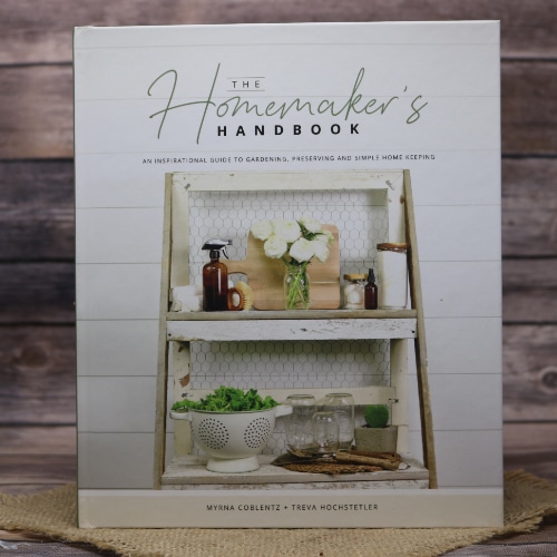 The cover of 'The Homemaker's Handbook' featuring an elegant shelf with cleaning supplies and home decor, set on a straw mat with a wooden background.
