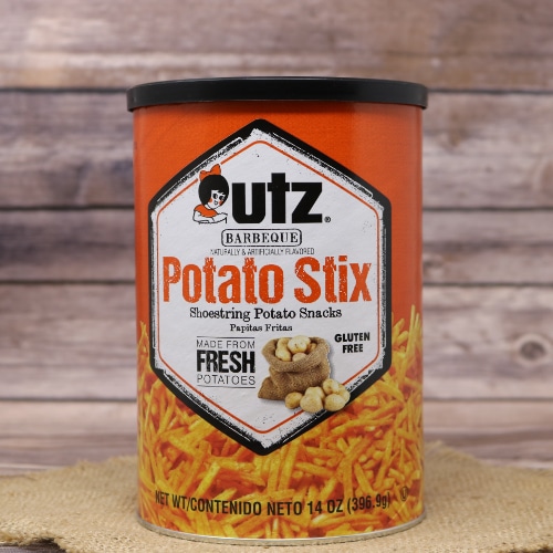 A cylindrical container of Utz Barbeque Potato Stix, showcasing the product against a straw mat and wooden background.