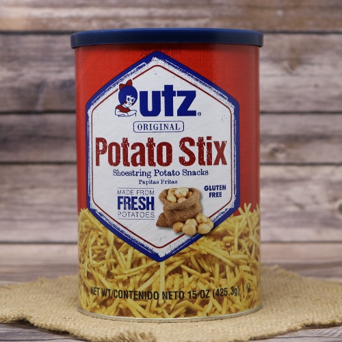 Canister of Utz Original Potato Stix in a red and blue design, displayed on a natural straw mat with a soft-focus wood panel backdrop.